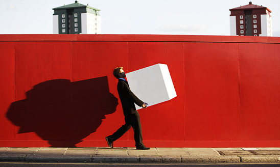 A man in a suit carries a big white box down a sidewalk in front of a red wall