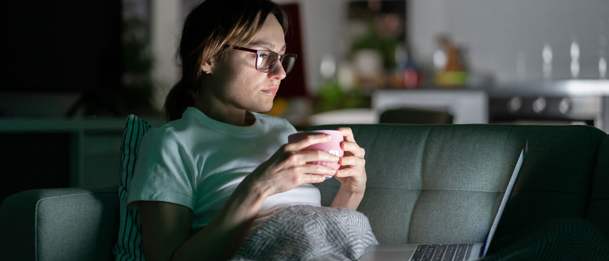 A woman sits on the couch in the glare of her laptop screen