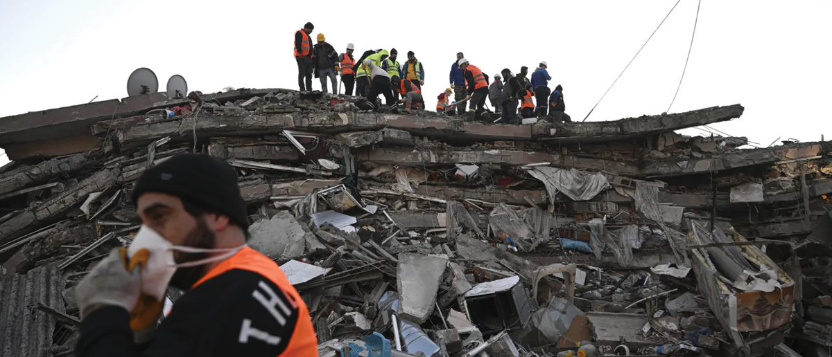Relief workers in brightly colored vests stand on a mound of rubble; satellite dishes and other debris suggest that it had previously been a building