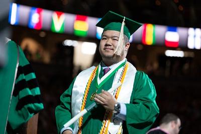 A graduate in regalia and honor cords receives his diploma in Spring 2023 commencement