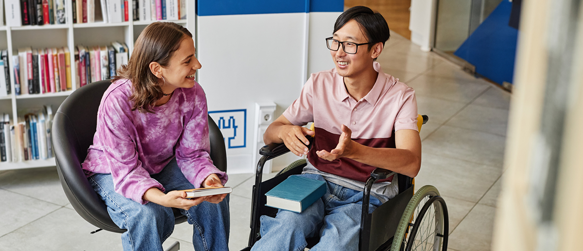 Two people with books in their laps sit and discuss, one sitting in a chair and one using a wheelchair